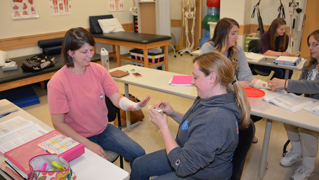 Two girls sitting in a chair bandaging a hand.